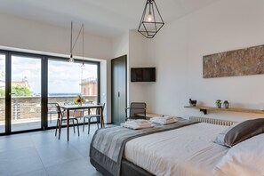 Une chambre moderne avec un grand lit, un bureau et une chaise, le tout près d’une porte-fenêtre coulissante donnant sur un balcon.