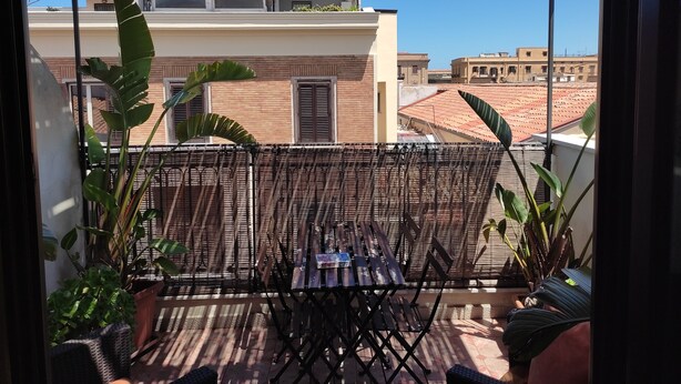A balcony with a wicker chair, potted plants, and a view of neighboring buildings. - Bright Penthouse With Terrace On Via Maqueda, In The Heart Of The City (Palermo)