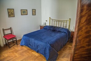 Une chambre avec un plancher en bois, un lit recouvert d’une couette bleue, une table de chevet en bois, une chaise avec un coussin rouge et deux tableaux encadrés accrochés au mur.