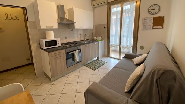 A compact kitchen with a microwave, stove, and sink, adjacent to a gray sofa and a sliding glass door leading to a balcony.