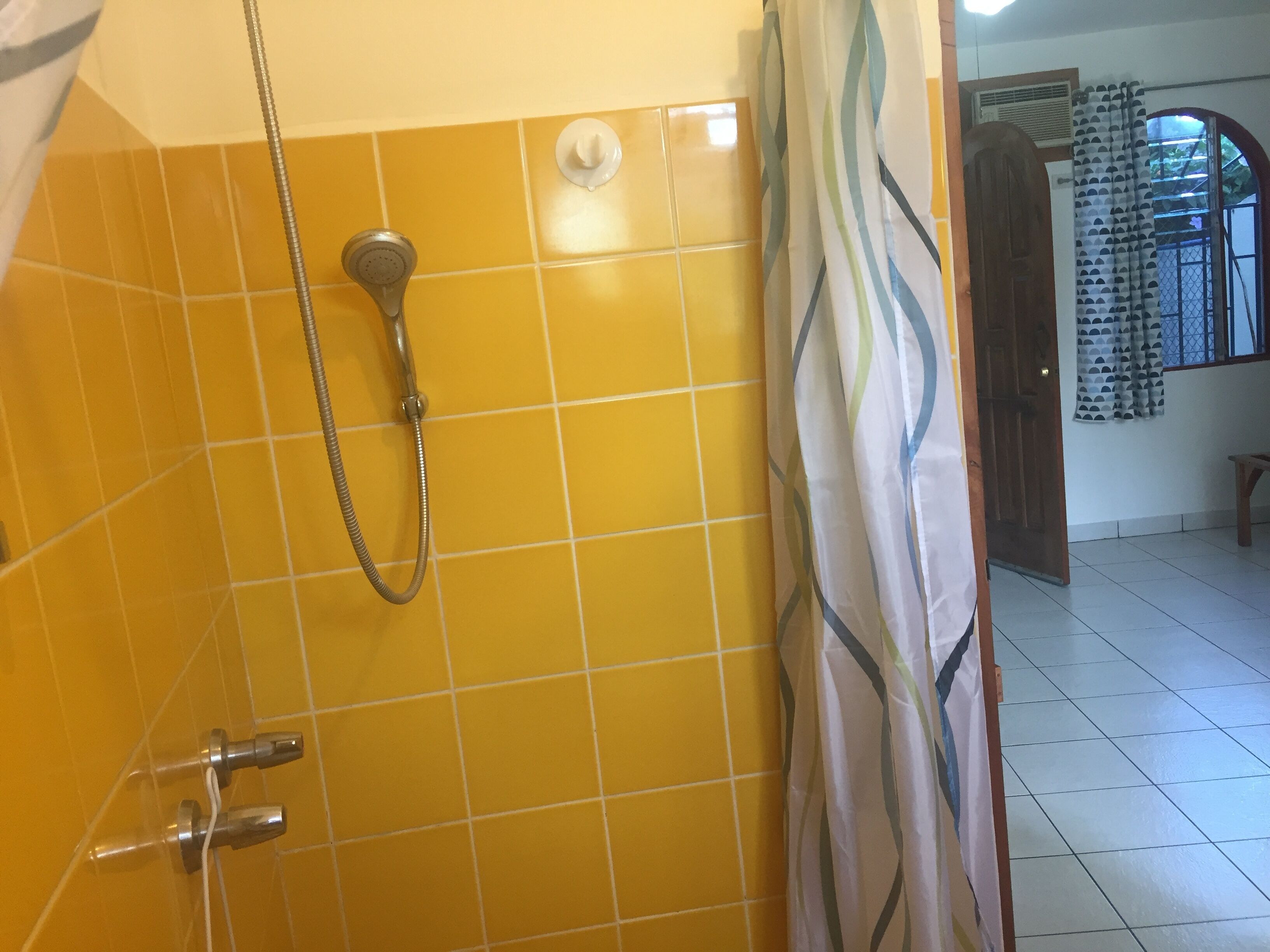 A bathroom with yellow tiled walls, a shower with a handheld hose, and a window with patterned curtains.