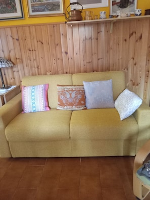 A sofa with patterned cushions, a wooden wall panel, and a shelf with decorative items.