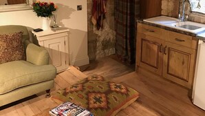 Un salon chaleureux avec un canapé, une table basse sur laquelle sont posés des magazines, une commode en bois et un coin cuisine avec un mur en pierre, des placards en bois et un évier.