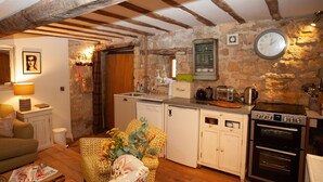 Une cuisine rustique aux murs de pierre, avec des poutres apparentes, un four encastré et un coin repas agrémenté d’une chaise et d’une table à motifs.