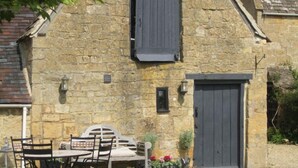Un bâtiment en pierre avec une porte en bois sombre, une petite fenêtre et un banc avec des chaises à l’extérieur.