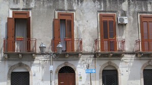 A building with three windows, each with brown shutters and a balcony with a metal railing. There is a street lamp and a sign with arrows pointing to the right.