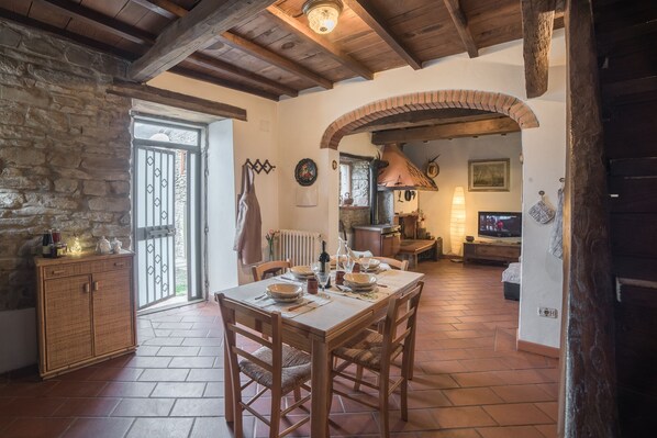 A rustic dining area with a stone wall, wooden ceiling beams, and a brick archway leading to a kitchen with a wood stove.