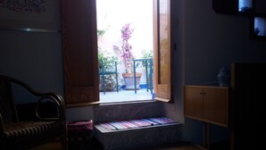 Una habitación con piso de madera, una silla de mimbre, un banco con una manta colorida, un armario de madera y vista a un jardín por una puerta abierta.