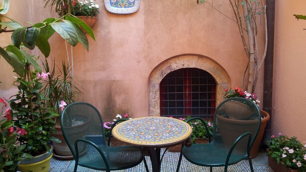 Un pequeño patio al aire libre con una mesa y dos sillas de cerámica, rodeado de plantas en macetas y un azulejo decorativo en la pared.