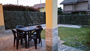 Une terrasse extérieure avec une table et des chaises en pierre, un mur en pierre et une zone herbeuse avec un tuyau d’arrosage.