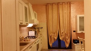 Une cuisine compacte avec des meubles blancs, un four à micro-ondes, une cuisinière et un évier. Il y a une fenêtre avec des rideaux et une petite table avec une imprimante.