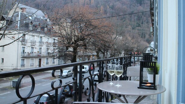 Ein Balkon mit Tisch und zwei Weingläsern, Blick auf eine Straße mit geparkten Autos und Gebäuden, mit Bergen im Hintergrund.