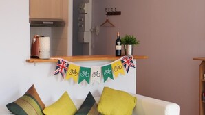 Ein modernes Wohnzimmer mit einem weißen Sofa, grünen und gelben Kissen, Holzboden und einer Küche im Hintergrund.