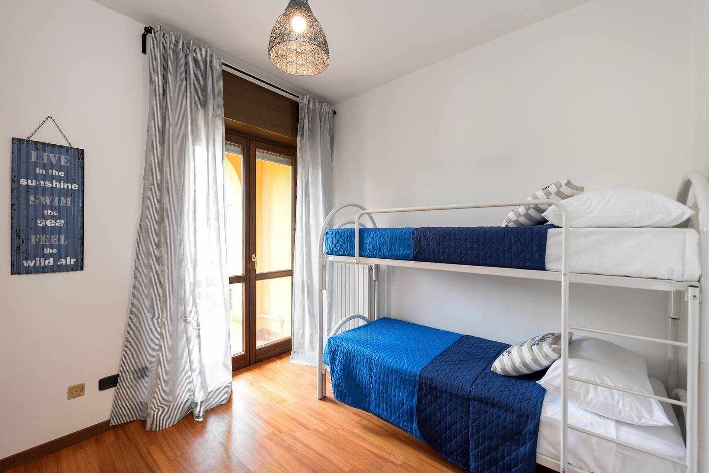 Een slaapkamer met een stapelbed, een houten vloer, witte muren, een raam met gordijnen en een hanglamp.