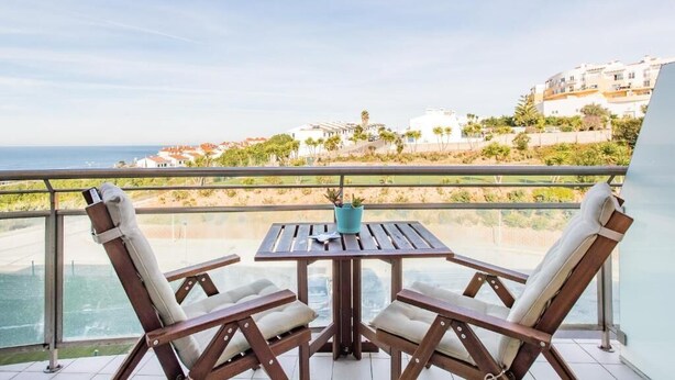 A balcony with two wooden chairs and a table, overlooking a coastal view with buildings and greenery.