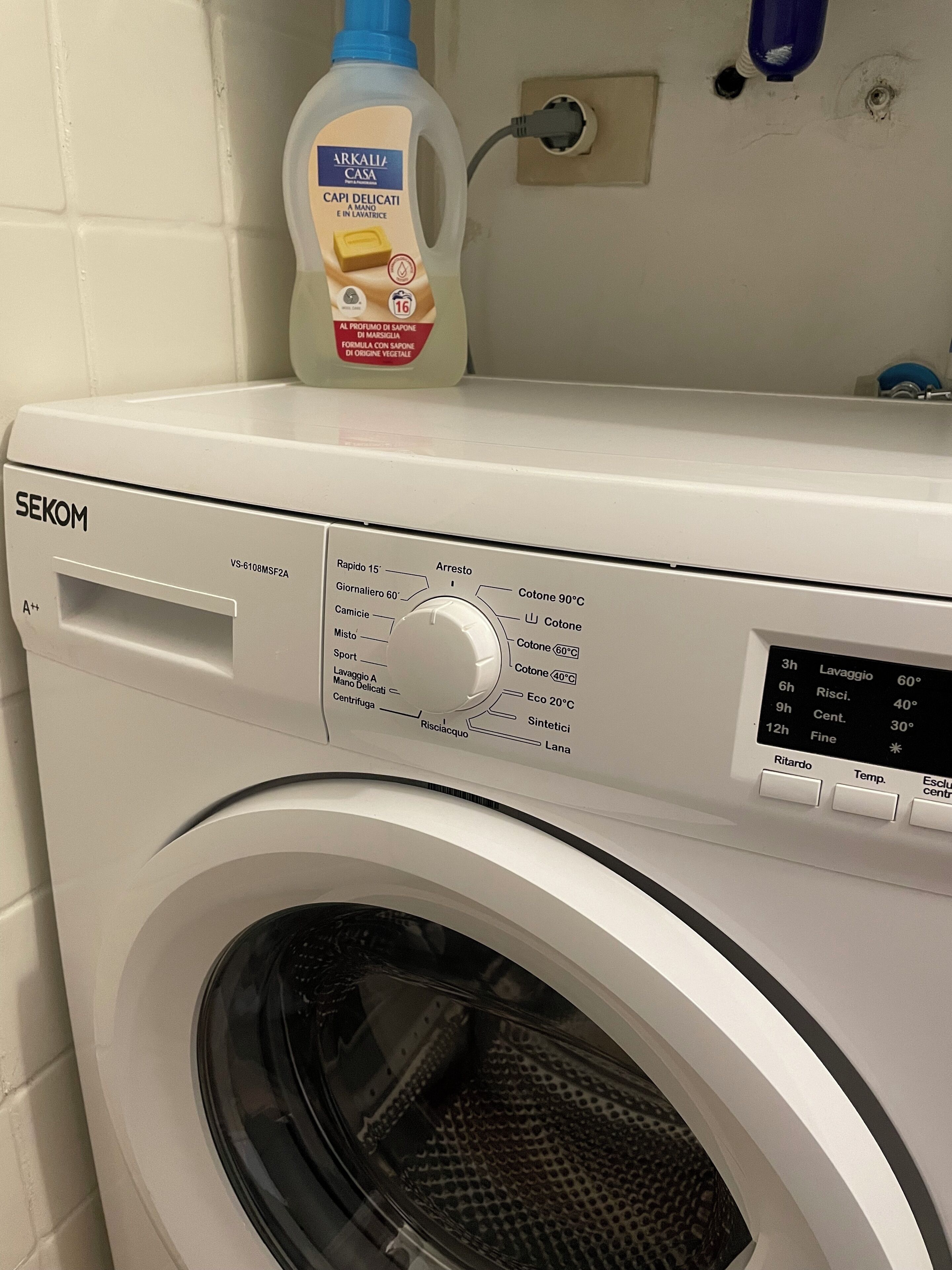 A front-loading washing machine with a control panel displaying various settings and a bottle of liquid soap on top.