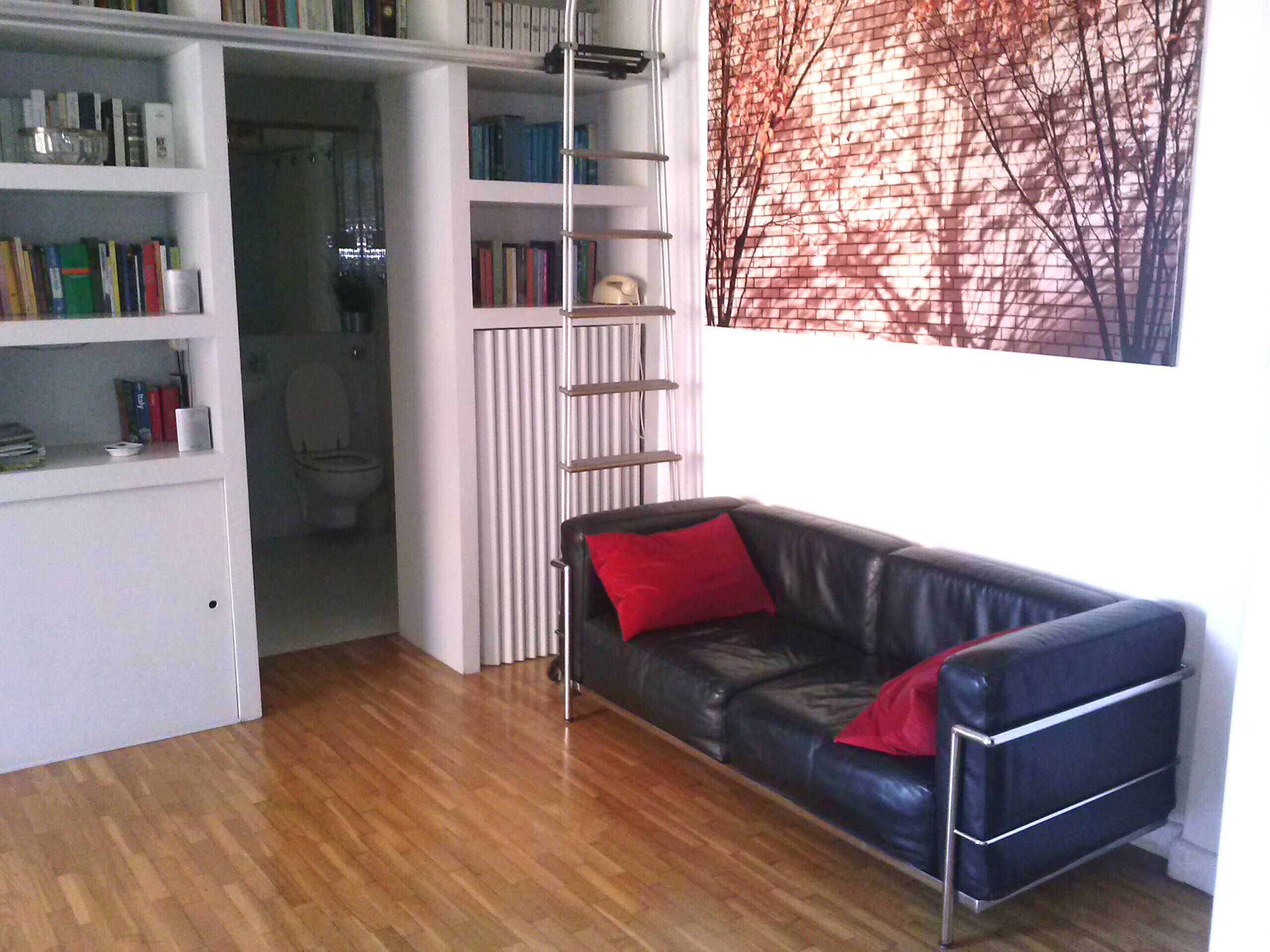 Una habitació amb un sofà de pell negra, una biblioteca i vista a una paret de maó.