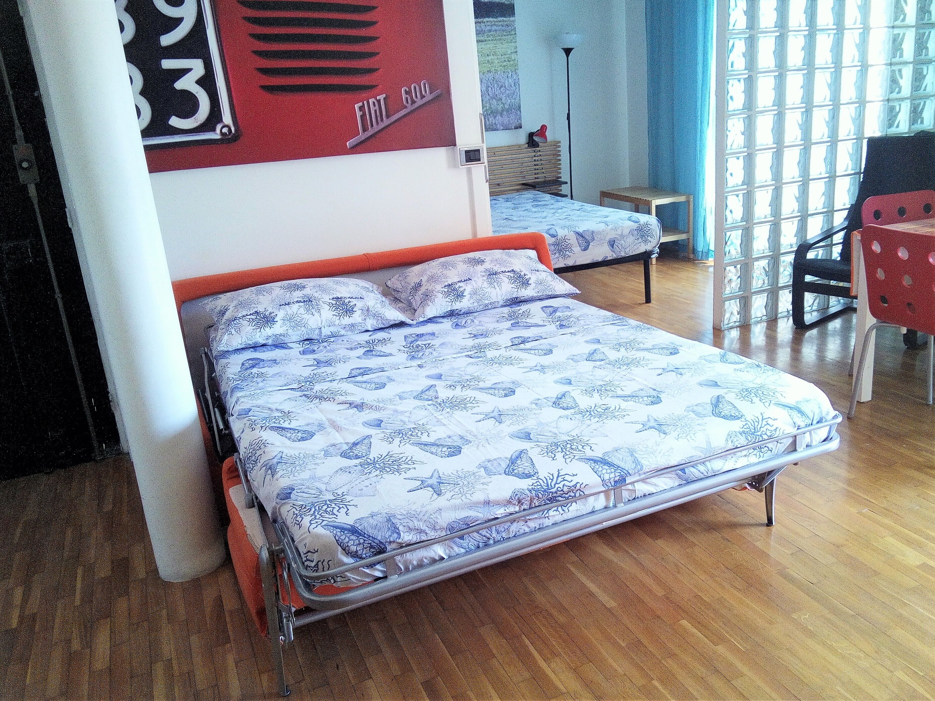 A single-bed room with a patterned bedspread, a wooden floor, and a red chair.