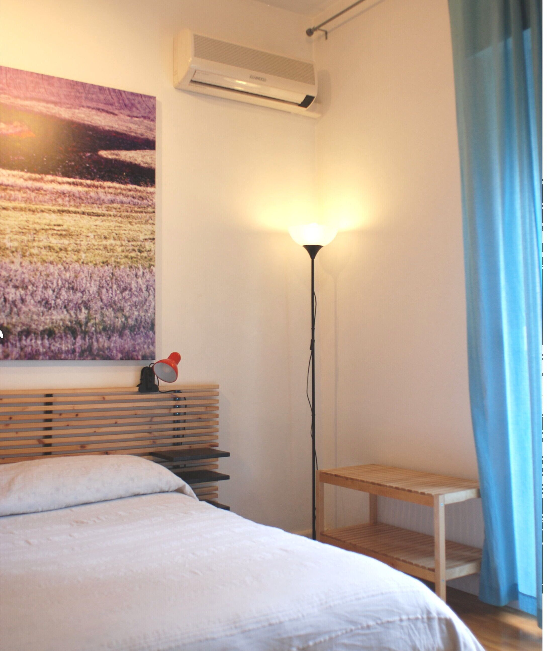 A bedroom with a bed, a bedside table, a lamp, a wall-mounted air conditioner, and a painting.