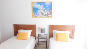 Ein Hotelzimmer mit zwei Betten, jeweils mit weißer Bettwäsche und einem gelben Kissen. Dazwischen steht ein kleiner Tisch mit einer Lampe. Darüber hängt ein gerahmtes Bild mit Blumen an der Wand.