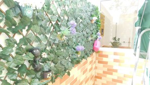 Eine Hausecke mit einer Ziegelwand und einer begrünten Wand. Auf der grünen Wand stehen Blumentöpfe und ein pinkfarbener Wasserkrug.