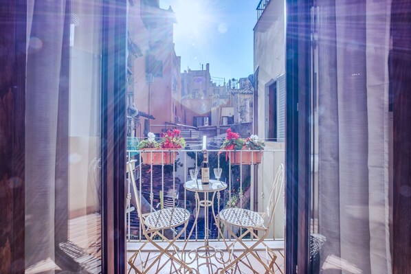 A balcony with two chairs and a table, overlooking a narrow street with buildings.