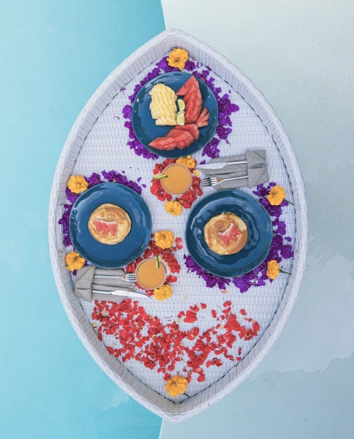 Una bandeja con tres platos azules, cada uno con una tajada de sandía, fruta y una bebida.