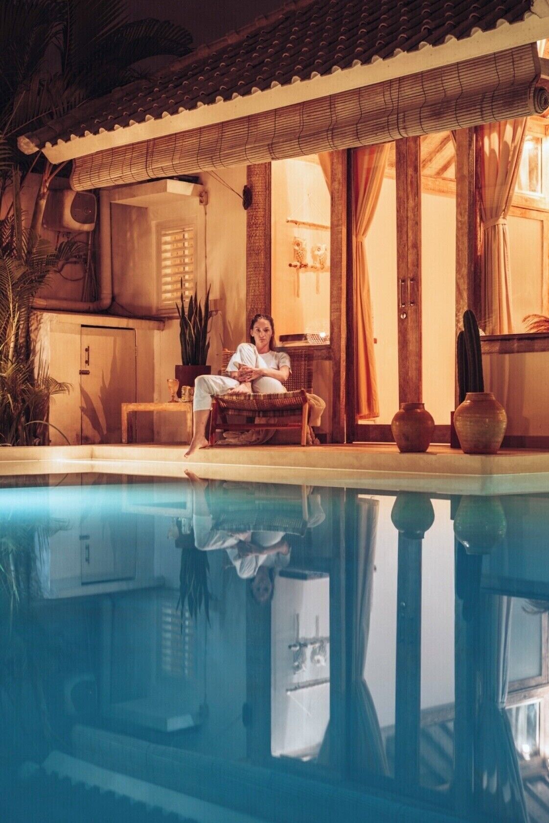 A person sitting by a pool with a traditional tiled roof and wooden furniture.
