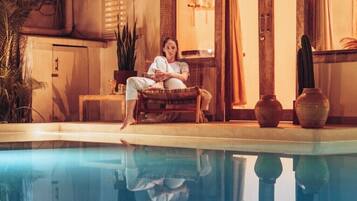 A person sitting by a pool with a traditional tiled roof and wooden furniture.