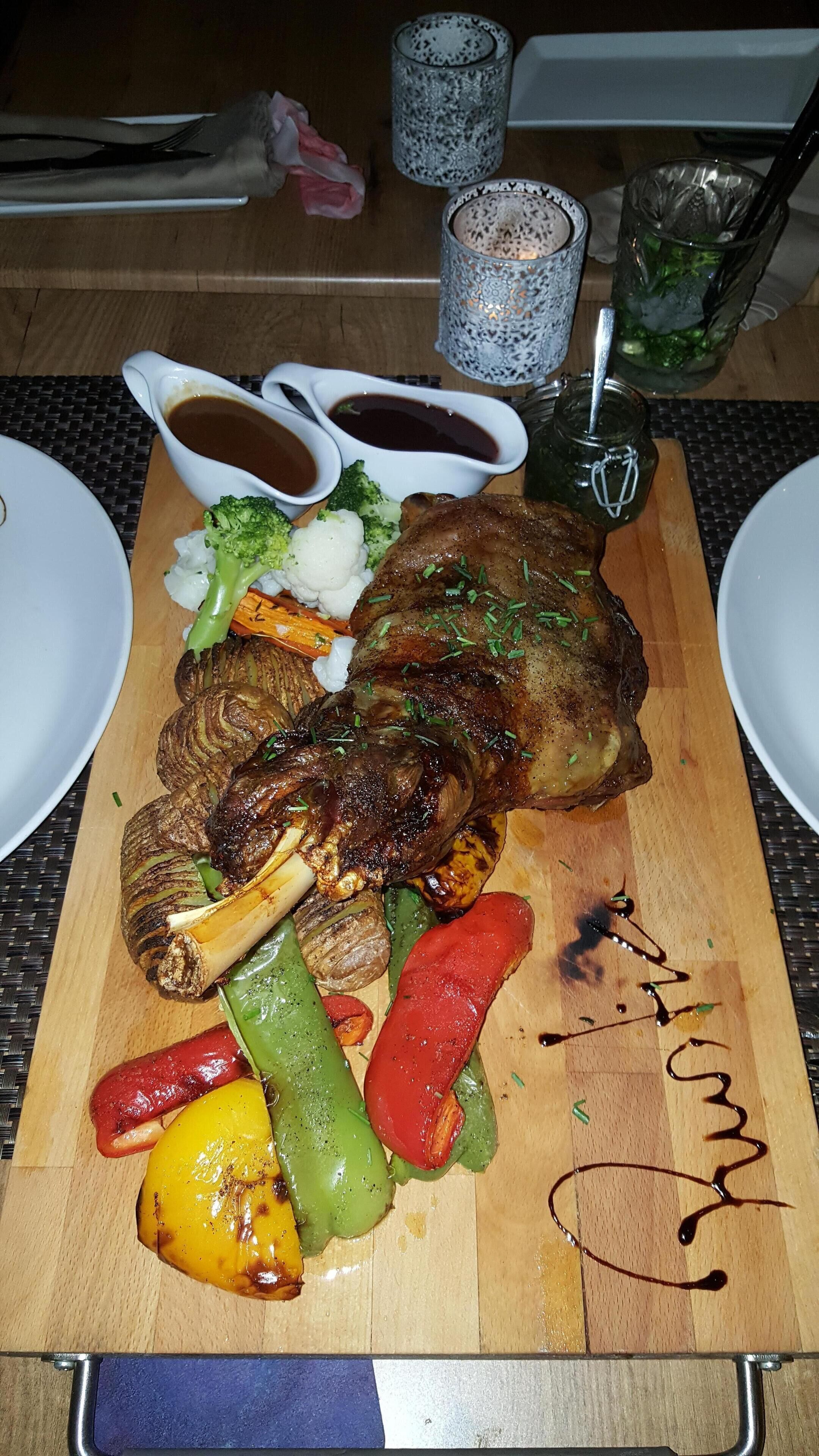 A wooden board with a roasted lamb leg, vegetables, and gravy bowls.