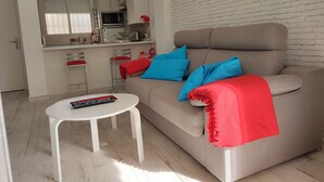 Un salón con un sofá color beige, cojines rojos y azules, una mesa de centro blanca y una zona de cocina con armarios blancos y un microondas.