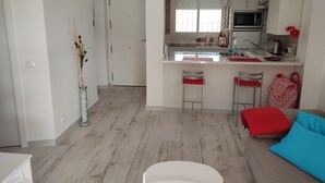 Una cocina moderna con microondas, pileta y taburetes de bar. Hay un comedor con una mesa blanca y un sofá con cojines rojos y azules.