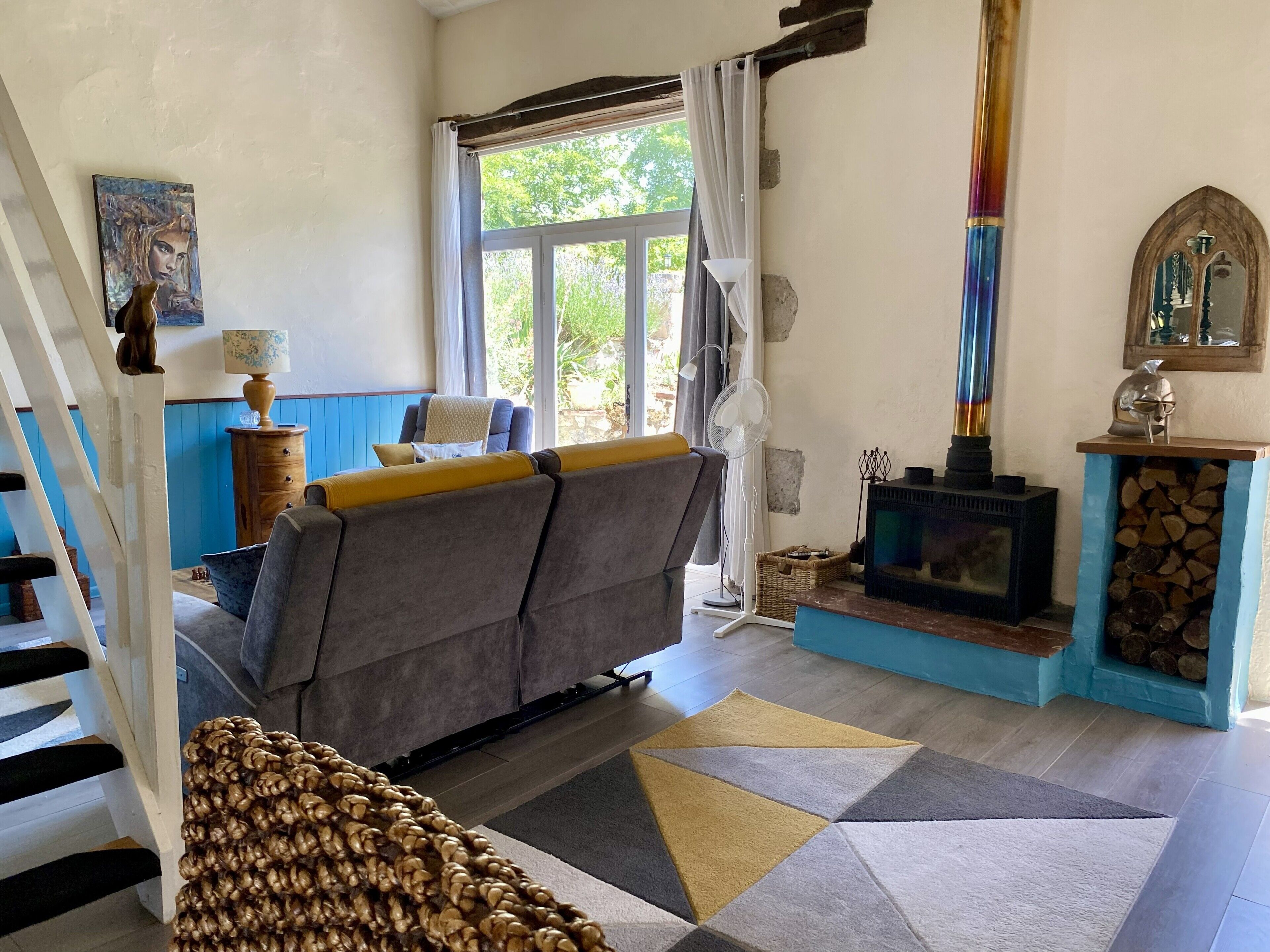 Un salon chaleureux avec un canapé, une cheminée, un escalier et une vue sur la verdure à l’extérieur.