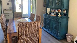 Een eetruimte met een houten tafel en gevlochten stoelen, een blauwgroene kast met decoratieve borden en een uitzicht door een glazen deur.