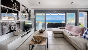 Un salon moderne doté d’un grand téléviseur, d’une table basse en bois, d’un canapé beige et offrant une vue sur la mer grâce à de grandes portes coulissantes.
