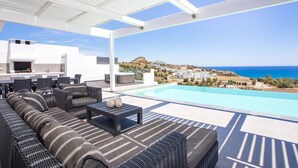 Un espace de détente extérieur moderne, doté d’une piscine, de sièges et offrant une vue sur la mer et les collines.