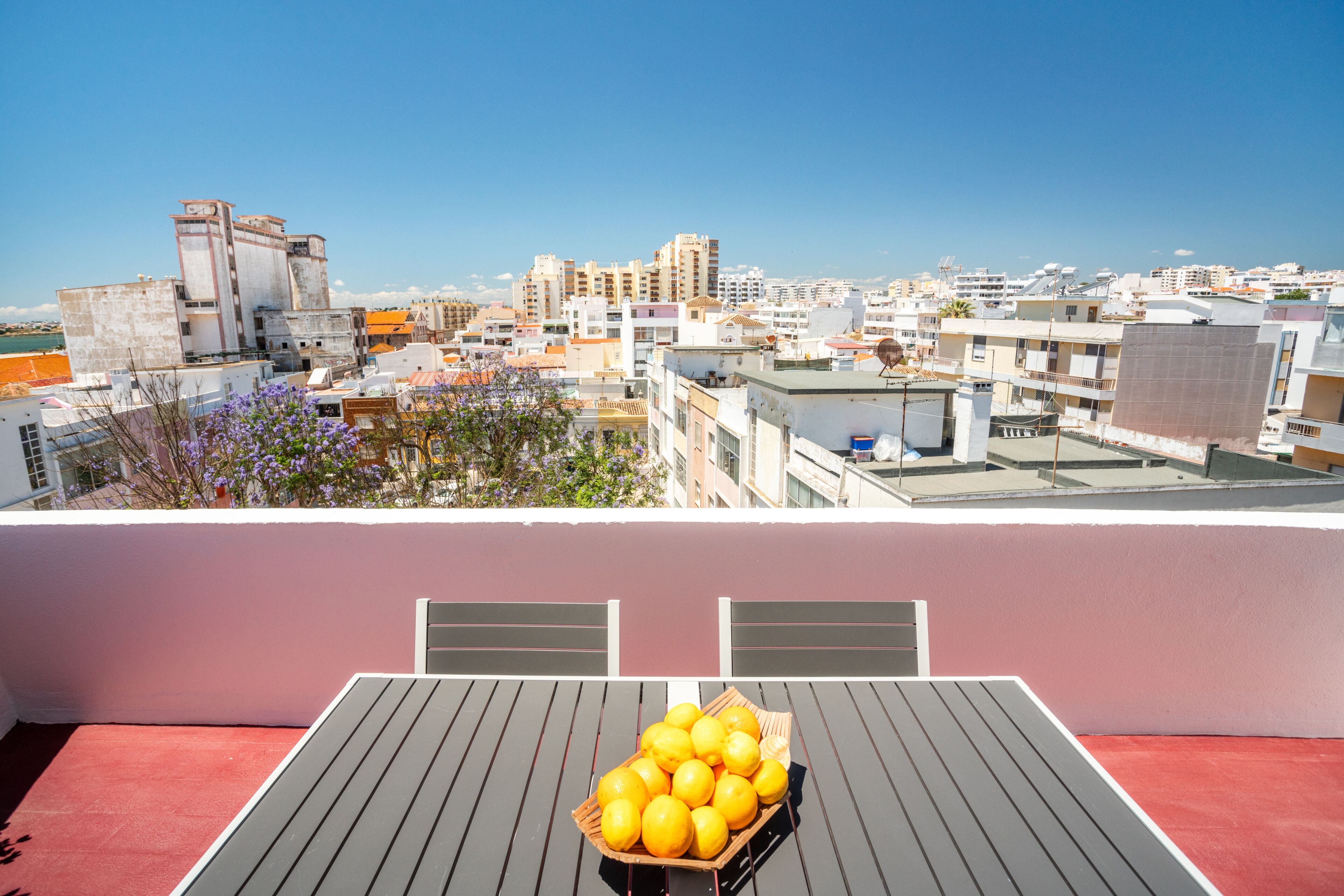 A rooftop terrace with a table and a basket of lemons, overlooking a cityscape with buildings and a clear sky.