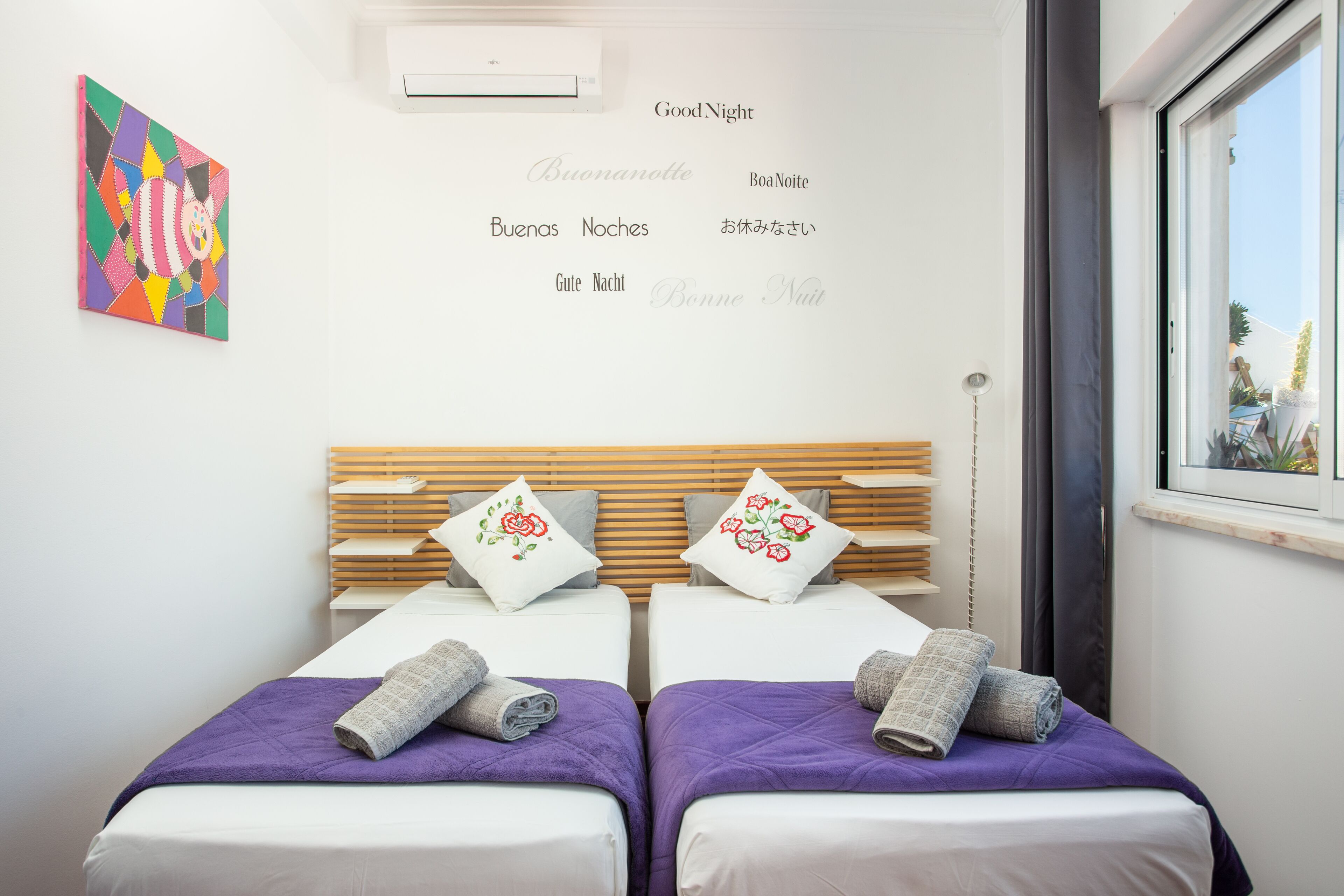 Ett hotellrum med två sängar, ett fönster, en väggmonterad luftkonditioneringsanläggning och ett färgstarkt konstverk.