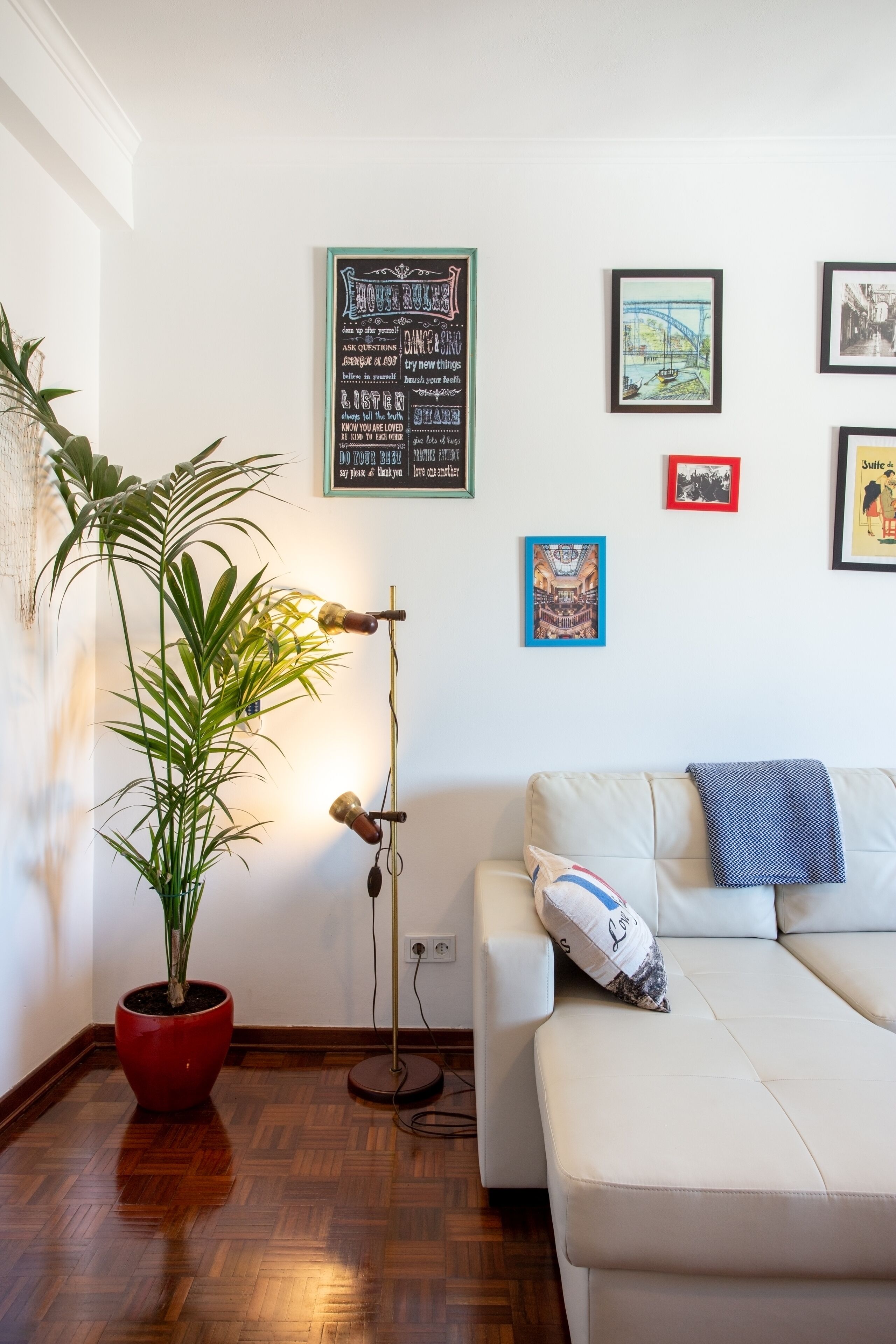 Una sala de estar con sofá, una planta en maceta roja, una lámpara de pie y cuadros en la pared.
