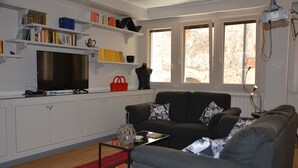 A living room with a grey sofa, a red rug, a coffee table, and a TV on a white cabinet. Shelves with books and decorative items are above the TV. - Large apartment with 2 bedrooms and two bathrooms in PALAZZO SAN MATTEO (Palermo)