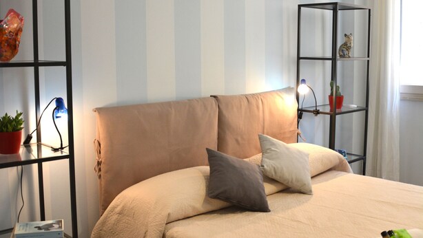 A neatly arranged bedroom with a bed, pillows, a shelf with a plant, and a window with curtains. - Large apartment with 2 bedrooms and two bathrooms in PALAZZO SAN MATTEO (Palermo)