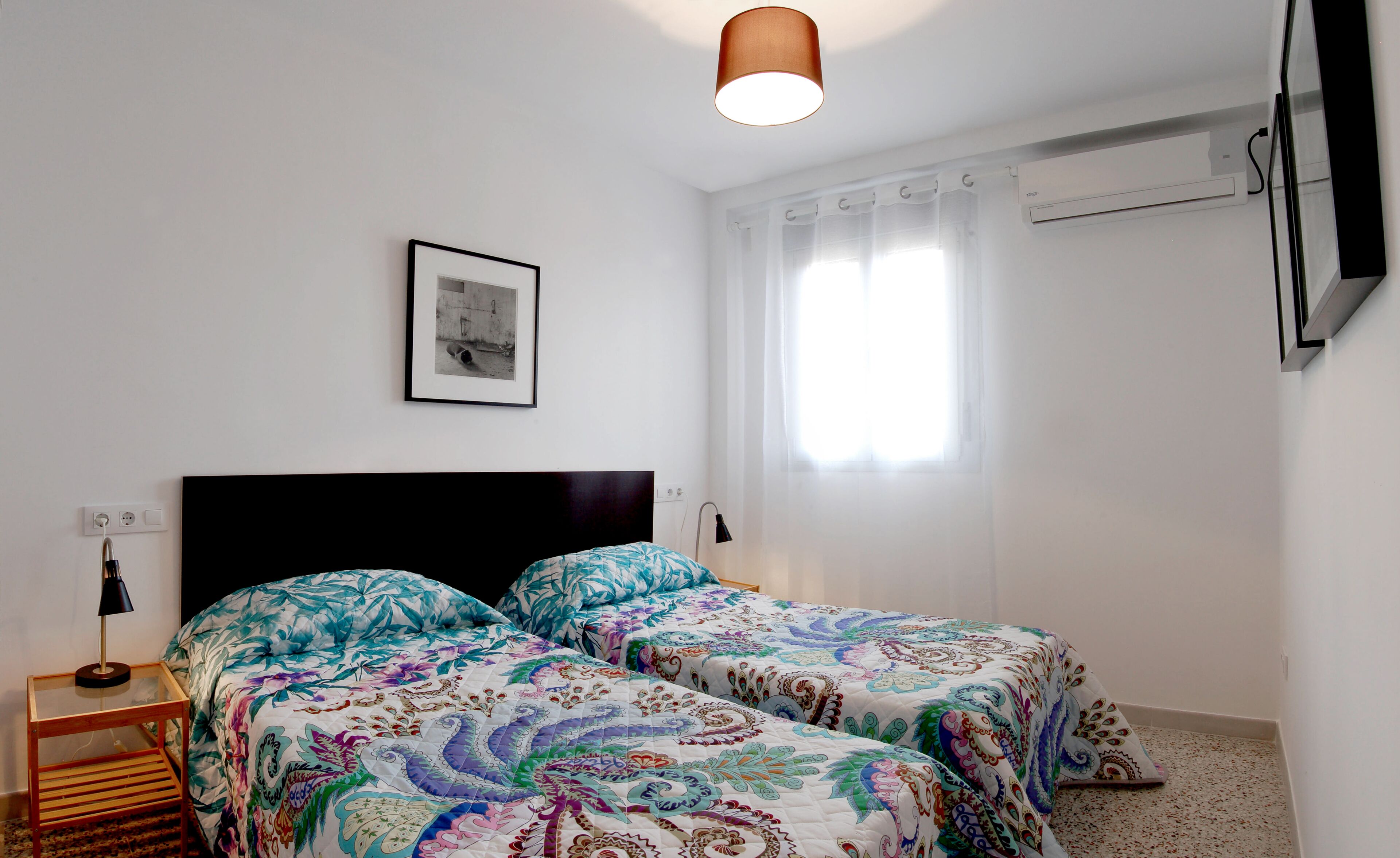 A bedroom with two beds, a framed picture on the wall, a ceiling light, and an air conditioning unit.