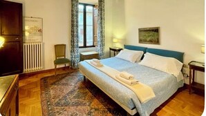A bedroom with a bed, a chair, a small table, a radiator, and a window with curtains.