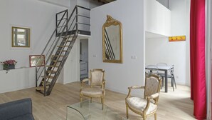 Een ruime kamer met een houten trap, antieke stoelen, een glazen tafel en een grote spiegel.