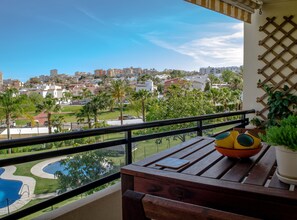 Uma varanda com mesa de madeira, planta em vaso e vista para área residencial com edifícios e piscina.