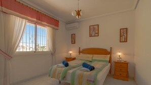 A bedroom with a bed, two bedside tables, a window with curtains, and wall-mounted air conditioning.