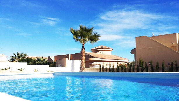 A swimming pool with clear blue water, surrounded by palm trees and a building with a tiled roof.