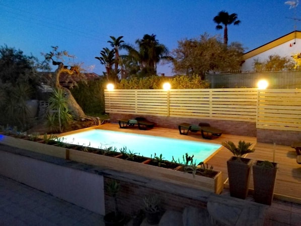 A swimming pool with a wooden deck, surrounded by plants and a fence.