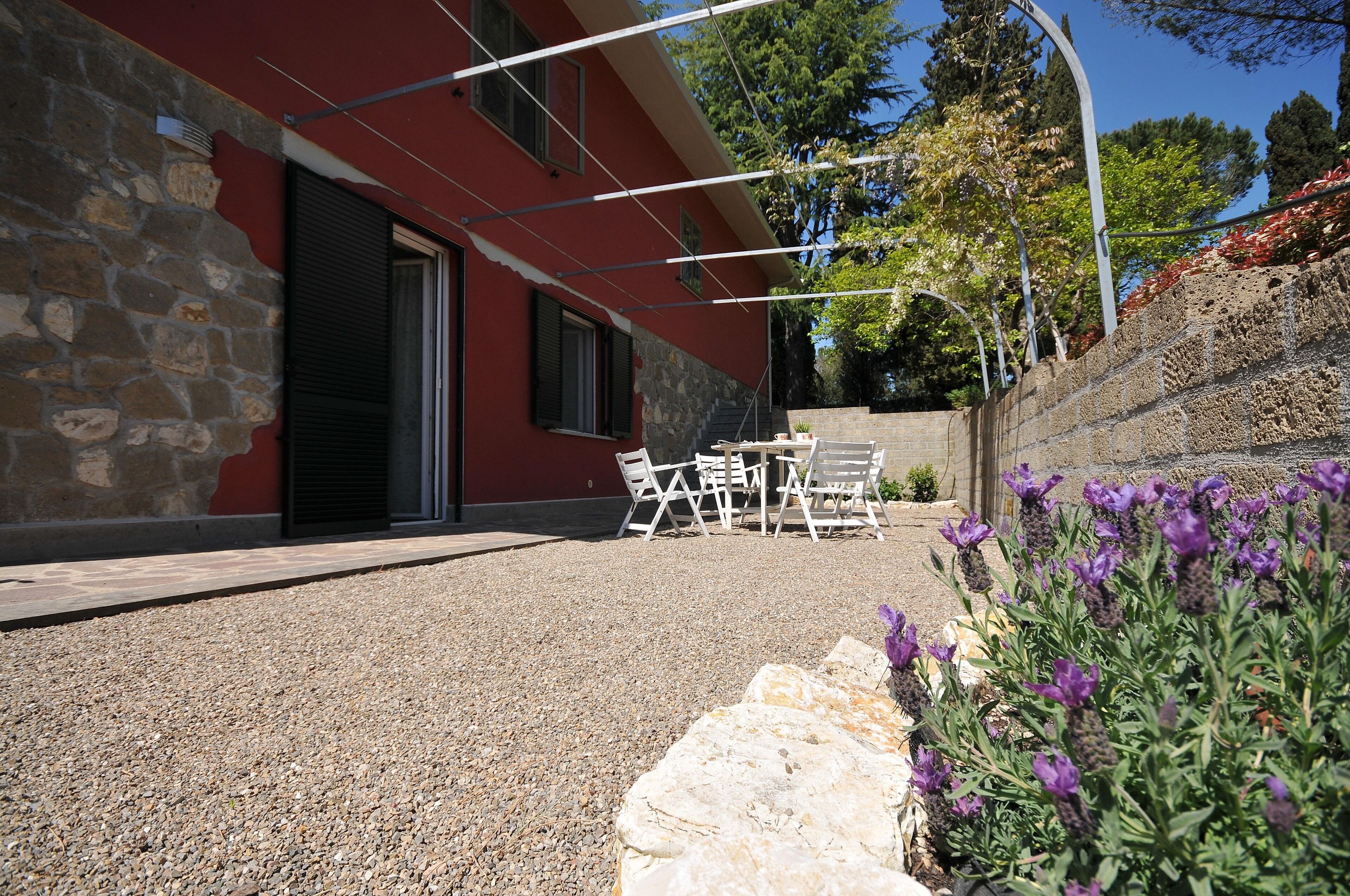 Un edificio con fachada de piedra y pared exterior roja. Cuenta con un patio con mesa y sillas, y un jardín con flores púrpuras.