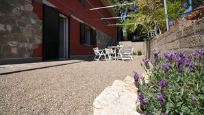 Een gebouw met een stenen gevel en een rode buitenmuur. Er is een terras met een tafel en stoelen, en een tuin met paarse bloemen.